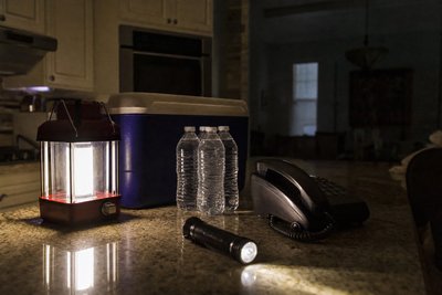 A kitchen counter at dusk lit by a battery lantern and flashlight, with a cooler, bottled water, and a corded phone nearby during a power outage